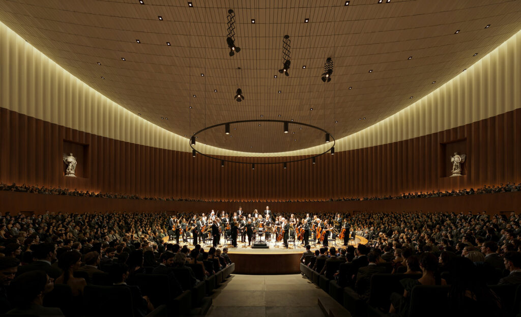 A large orchestra on a luminous stage, encircled by audience; refined wooden walls and an arching ceiling define the acoustic space.